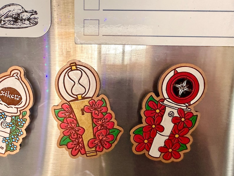 May include: Wooden magnets with a floral design. One magnet features a hourglass shape, and the other has a compass design. Both magnets are decorated with red flowers and green leaves.