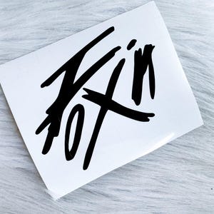 Puede incluir: Una calcoman&iacute;a rectangular blanca con un dise&ntilde;o gr&aacute;fico negro audaz y de estilo dibujado a mano. La obra consiste en l&iacute;neas gruesas y expresivas similares a pinceladas que forman una forma abstracta y estilizada en la superficie del adhesivo.