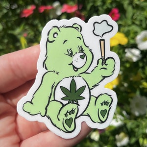 May include: Green Care Bear sticker with a marijuana leaf on its chest and a joint in its hand. The bear is smiling and has a cloud of smoke above its head.