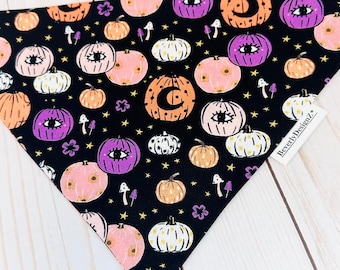 May include: Black bandana with a repeating pattern of orange, pink, and white pumpkins with purple eyes and a crescent moon on a black background. The bandana has the text "Beverly Designz" on the bottom right corner.
