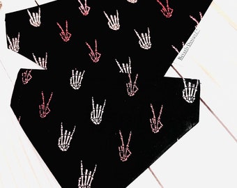 May include: Two black dog bandanas with a repeating pattern of pink and white skeleton hand gestures. One bandana is folded over the other, displaying the design. The bandanas are on a white wooden surface.
