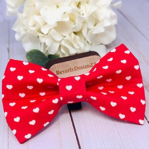 May include: Red bow tie with white hearts. The bow tie is made of fabric and has a small black strap in the back.