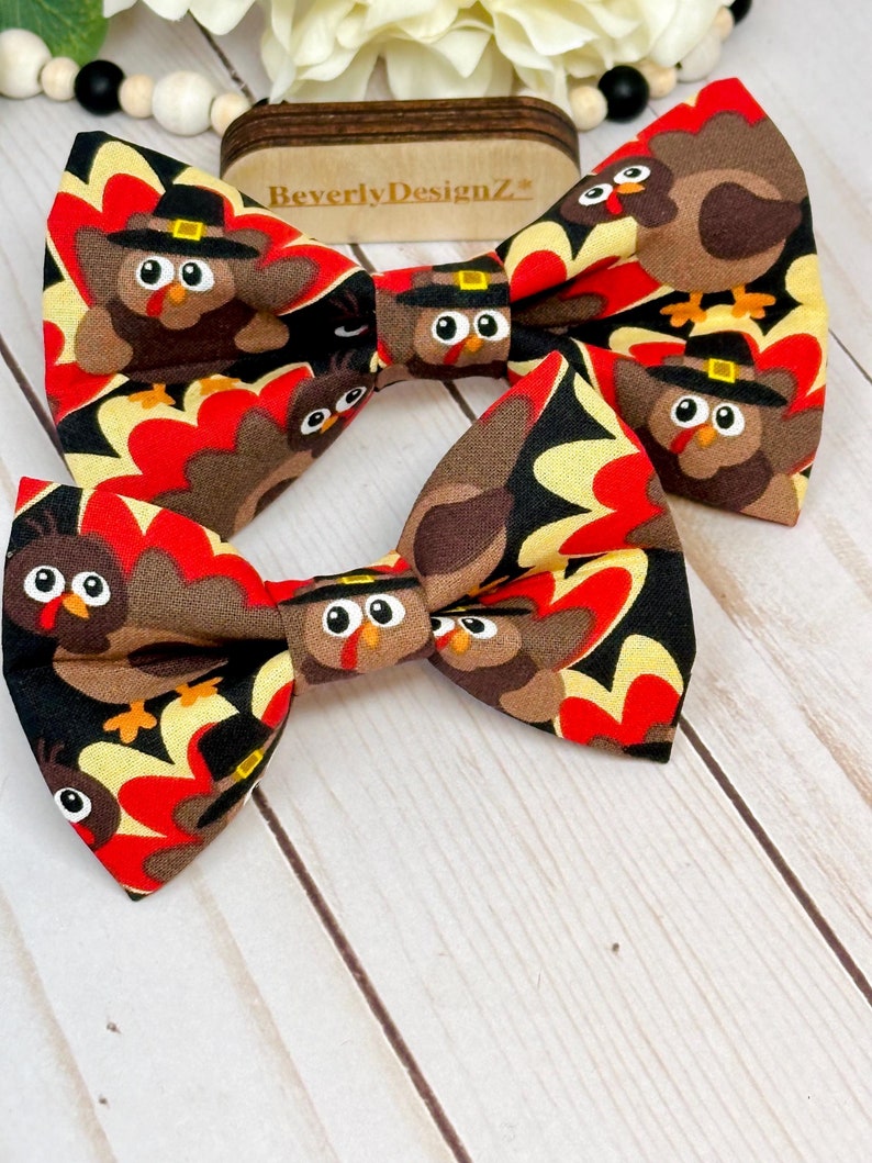 May include: Two Thanksgiving-themed bow ties. The fabric features a repeating pattern of cartoon turkeys wearing pilgrim hats, set against a backdrop of red, yellow, and black shapes. The bow ties are on a white wooden surface.