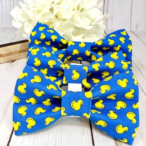May include: Two blue bow ties with a yellow rubber ducky pattern. The bow ties are made of fabric and have a white adjustable strap.
