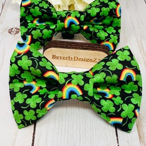 May include: Two green shamrock and rainbow bow ties on a wooden surface. The bow ties are made of fabric and have a black background.