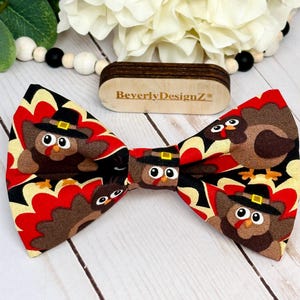 May include: A Thanksgiving-themed bow tie featuring a repeating pattern of cartoon turkeys wearing pilgrim hats. The bow tie has a black, red, and yellow background. The brand name "BeverlyDesignZ" is visible on a wooden tag.