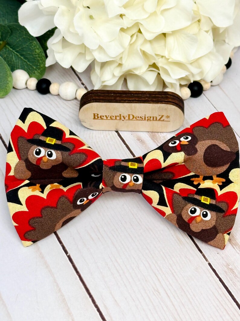 May include: A Thanksgiving-themed bow tie featuring a repeating pattern of cartoon turkeys wearing pilgrim hats. The bow tie has a black, red, and yellow background. The brand name "BeverlyDesignZ" is visible on a wooden tag.