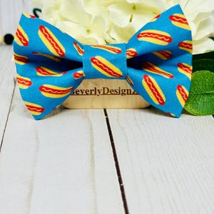 May include: A blue bow tie with a repeating pattern of yellow and red hot dogs. The bow tie is centered on a wooden block with the text "BeverlyDesignz". White flowers and green leaves are in the background.
