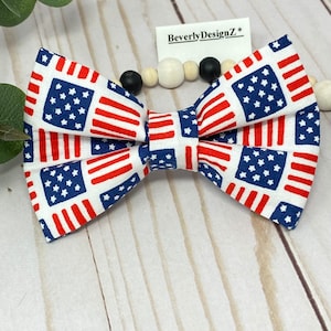 May include: A red, white, and blue bow tie with a pattern of American flags. The bow tie is made of fabric and has a white background. The bow tie is tied with a white ribbon. The bow tie is on a white wooden background. The text "BeverlyDesignZ*" is on a white tag.