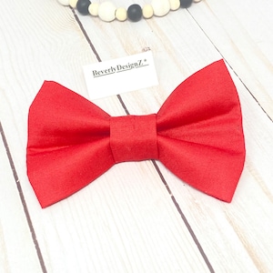 May include: A bright red bow tie with a classic shape, lying on a light wood surface. The bow tie is the focal point, with a tag that reads "BeverlyDesignZ*". A decorative wooden bead garland with black and white beads is in the background.