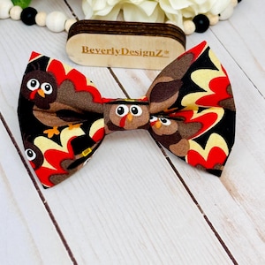 May include: A handmade bow tie featuring a turkey design with brown, red, black, and yellow colors. The bow tie has a repeating pattern of cartoon turkeys. The wooden tag in the background has the text "BeverlyDesignZ*".