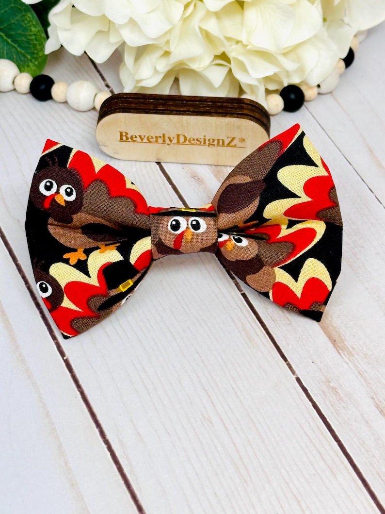 May include: A handmade bow tie featuring a turkey design with brown, red, black, and yellow colors. The bow tie has a repeating pattern of cartoon turkeys. The wooden tag in the background has the text "BeverlyDesignZ*".