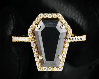 May include: A gold ring with a black diamond shaped like a coffin. The diamond is surrounded by smaller diamonds.