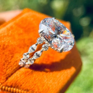 Puede incluir: Primer plano de un anillo de plata con un gran diamante de forma ovalada. El anillo está engastado con diamantes más pequeños en la banda. El anillo está sobre una superficie de terciopelo naranja brillante.