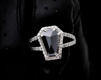 May include: A black diamond ring with a coffin-shaped setting. The ring is set in white gold and features a halo of smaller diamonds.