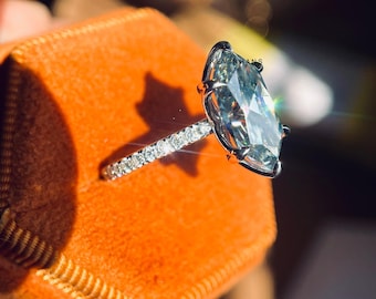 May include: A silver ring with a large, clear oval-shaped diamond set in a pave setting. The ring is in a small orange velvet box. The box has the text "KEVIN HANS" on the side.