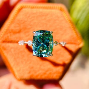 Puede incluir: Un anillo de compromiso con una piedra preciosa verde de corte cojín, una banda de plata y pequeñas piedras de acento. El anillo está en una pequeña caja de terciopelo naranja.