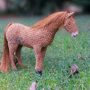 Puede incluir: Un juguete de caballo de crochet marrón con una larga melena y cola. El caballo está de pie en un campo de hierba verde.