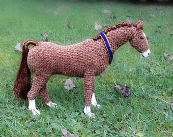 May include: A brown crocheted horse with a blue and gold ribbon around its neck. The horse is standing on a green grassy surface.