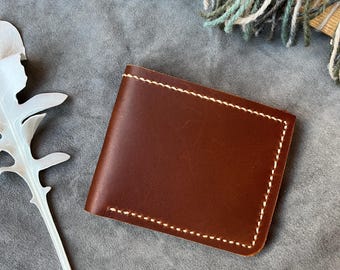 May include: A brown leather wallet with white stitching around the edges. The wallet is rectangular and appears to be bi-fold. The leather has a smooth texture. The background is a muted gray.