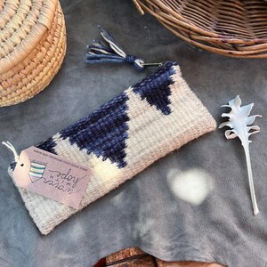 May include: A woven zippered pouch with a geometric pattern in shades of blue and white. The pouch is lying on a gray surface with a tag that reads "shop" and "himo" with a small woven design.