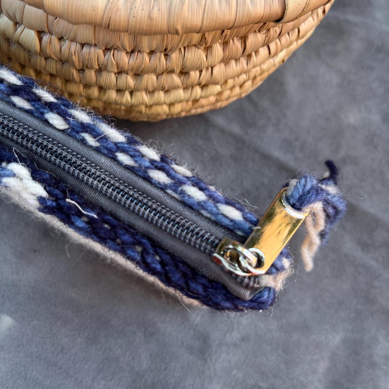 May include: A close-up of a zipper on a small, woven bag. The zipper is made of metal and has a gold finish. The bag is made of white and blue yarn.