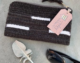 May include: A black and white woven clutch with a zipper closure and a tassel. The clutch has a tag that says "woven with hope" and "unique woven goods empowering rural women".
