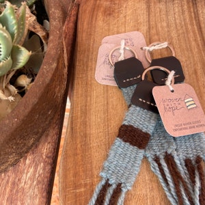 May include: Two blue and brown woven keychains with leather tassels and silver key rings. The keychains have a tag that reads "woven with hope" and "Unique woven goods empowering Arab women".