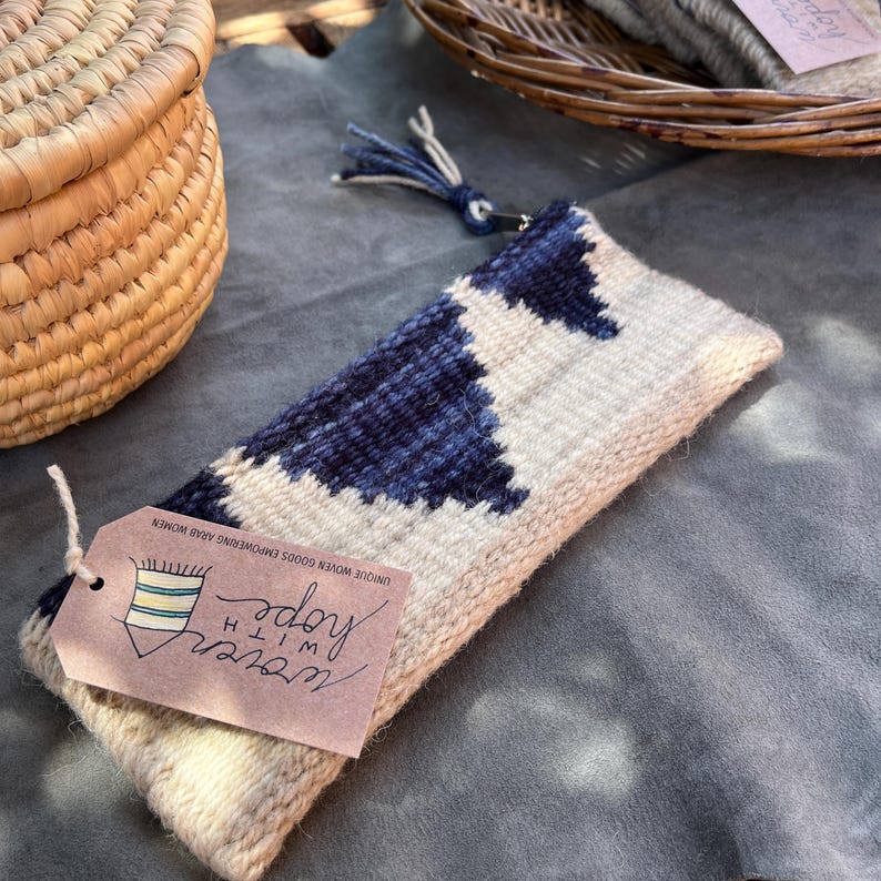 May include: A white and blue woven pouch with a zipper closure and a tassel. The pouch has a tag attached that says "woven with hope" and "unique woven goods empowering rural women".