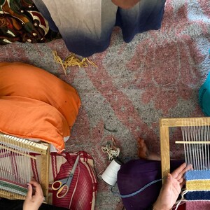 May include: Two people are sitting on the floor, each using a wooden loom to weave a small rug. One rug is pink, blue, yellow, and black. The other rug is green and white.