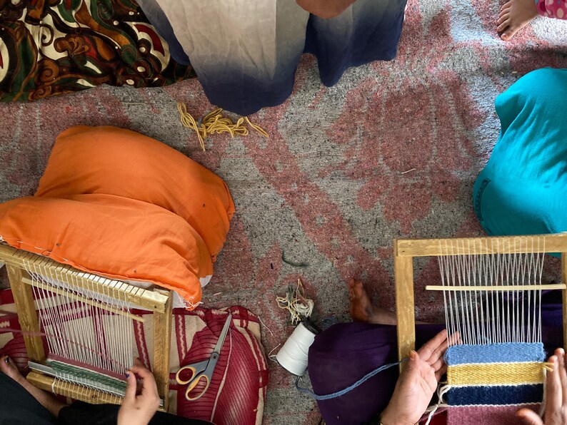 May include: Two people are sitting on the floor, each using a wooden loom to weave a small rug. One rug is pink, blue, yellow, and black. The other rug is green and white.