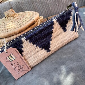 May include: A woven zippered pouch with a geometric pattern in shades of beige and dark blue. The pouch has a tassel and a tag that reads "I'm  day'  fashion" and "handmade woven goods empower women".