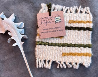 May include: A woven coaster with a tag that reads "woven with hope". The coaster is cream-colored with horizontal stripes of olive green and gold. The tag also says "Unique woven goods empowering Arab women."