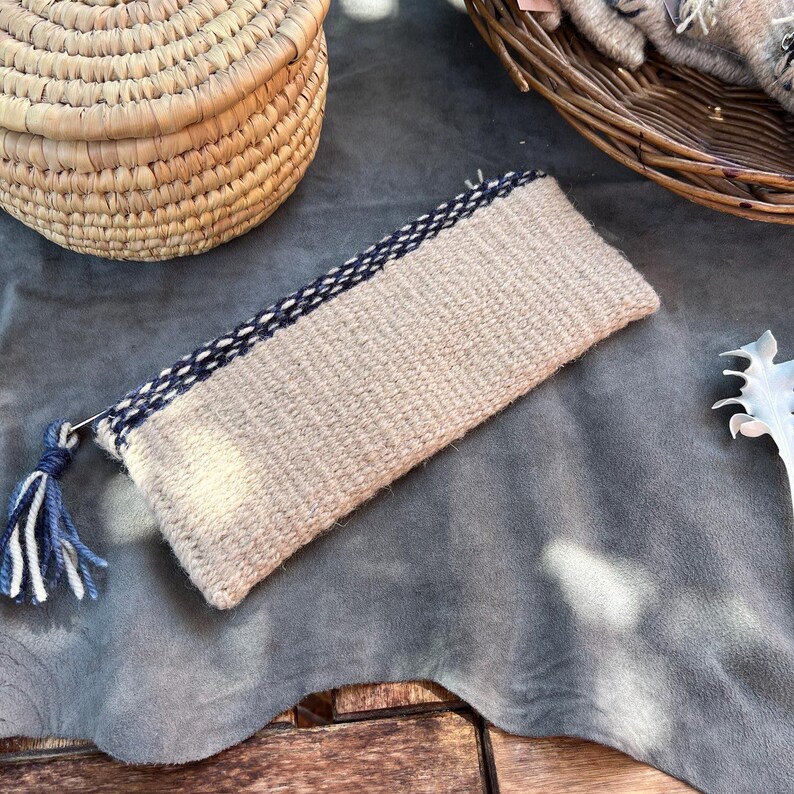 May include: A woven beige clutch with a black and white trim and a blue tassel. The clutch is lying on a gray surface.