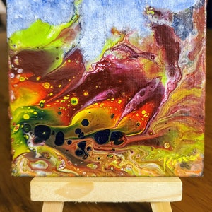 May include: Abstract painting on canvas with swirling colors of red, yellow, green, and blue. The painting is on a small easel.