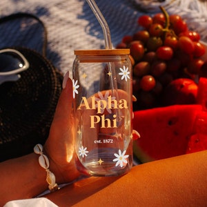 Peut inclure: Un gobelet en verre transparent avec un couvercle en bambou et une paille. Le gobelet est décoré d'un motif de marguerites blanches et du texte "Alpha Phi est. 1872" en lettres dorées.