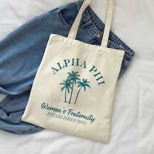 May include: A white canvas tote bag with a teal green design. The design features palm trees and the text "Alpha Phi Women's Fraternity Established 1872".
