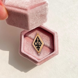 Puede incluir: Un anillo de oro en forma de diamante con una incrustación de esmalte negro que presenta un icono de espada y las letras griegas KA y O, presentado en un joyero hexagonal de terciopelo rosa con las letras KA grabadas en oro.