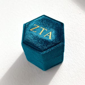 May include: A teal velvet hexagonal ring box with gold lettering spelling out "ZTA" on the lid. The box is closed and sits on a white surface. The box has a textured appearance.
