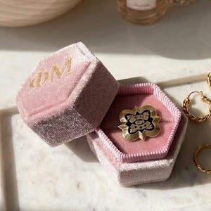 Könnte beinhalten: Eine rosa Samt-Ringbox mit goldenen Buchstaben, geöffnet, um einen goldenen Ring mit einem schwarz-goldenen Design zu enthüllen. Mehrere goldene Ringe und Creolen sind auf einem weißen Marmortablett angeordnet, zusammen mit Parfümflaschen.