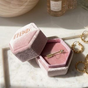 May include: A gold arrow ring with a small diamond is displayed in a pink velvet jewellery box. The box is open, revealing the ring. Gold hoop earrings and other gold rings are on a white marble tray.