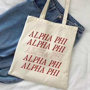 May include: A white canvas tote bag with the text "ALPHA PHI" repeated five times in red and pink. The bag is on top of a pair of blue jeans.