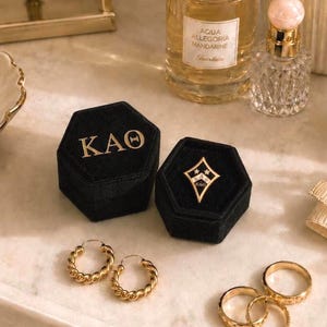 Könnte beinhalten: Zwei sechseckige Schmuckkästchen aus schwarzem Samt, eines mit den goldenen griechischen Buchstaben KAO und das andere mit einer goldenen rautenförmigen Anstecknadel mit KAO-Text. Dazu gehören goldene gedrehte Creolen und zwei strukturierte Ringe.