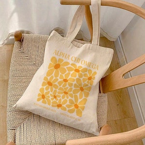 May include: A cream-colored tote bag with the text "ALPHA CHI OMEGA" and a pattern of yellow flowers. The bag has two handles and is resting on a woven chair. The text "ESTABLISHED 1885 GREENCASTLE IN" is printed below the flower pattern.