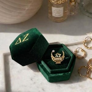 May include: A gold ring with a diamond and the letters "ΔZ" inside a dark green velvet ring box. The box is open, revealing the ring. Gold hoop earrings and other gold rings are also visible.