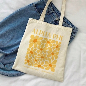 May include: A white canvas tote bag with the text "Alpha Phi" in black letters and a design of yellow daisies with orange centers. The text "Established 1872 Syracuse, NY" is printed below the daisies.