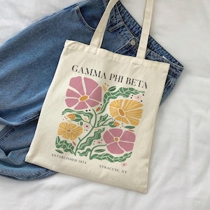 Peut inclure: Un sac fourre-tout crème avec un motif floral rose et jaune, et des feuilles vertes. Le texte "GAMMA PHI BETA" est imprimé au-dessus des fleurs, avec "ESTABLISHED 1874 SYRACUSE, NY" en dessous. Le sac est posé sur un jean bleu.