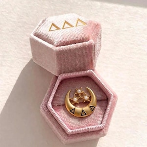 May include: A gold crescent moon brooch featuring three small stars with white pearls and black triangular accents, resting inside a hexagonal light pink velvet jewelry box with three gold triangle symbols embossed on the lid.