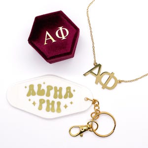 以下が含まれることがあります： 「AΦ」と書かれたチャームが付いたゴールドネックレスと、「ALPHA PHI」と金色で書かれた白いキーホルダーとゴールドのキーリング