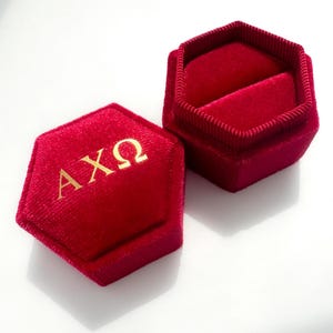 Op de afbeelding: Een rode fluwelen ringdoos met een zeshoekige vorm. De doos is open en toont een pluche interieur. Op het deksel staan de Griekse letters &Alpha;&Chi;&Omega; in goudkleurige letters. Ontworpen voor een ring.